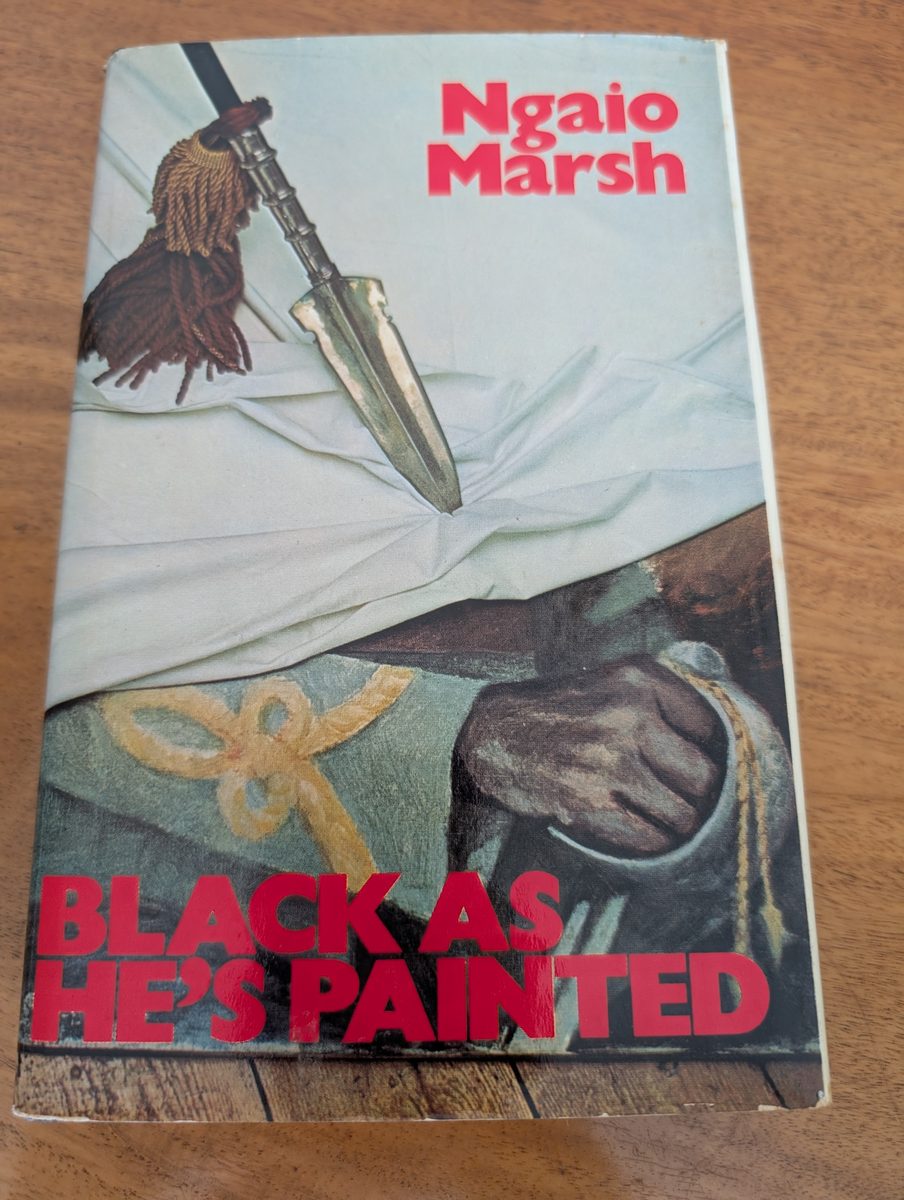 A photograph of the front cover of the 1974 UK hardback first edition of Ngaio Marsh's Black As He's Painted. The book is in good condition, with a dust jacket showing some light wear and creasing.