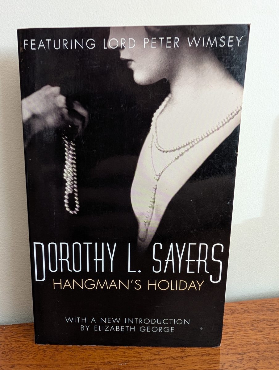The front cover of the 1974 New English Library paperback edition of Hangman's Holiday by Dorothy L. Sayers. The black cover features a close-up monochrome photograph of a woman in evening wear, her face in profile and turned away, wearing layered pearl and diamond necklaces and holding a strand of pearls in her hand. 'FEATURING LORD PETER WIMSEY' is printed in white at the top, 'DOROTHY L. SAYERS / HANGMAN'S HOLIDAY' in large white type in the centre, and 'WITH A NEW INTRODUCTION BY ELIZABETH GEORGE' at the bottom.