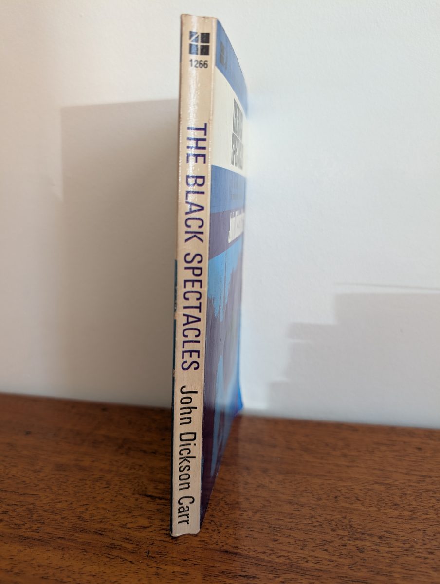 The spine of the 1965 Four Square Edition paperback of The Black Spectacles by John Dickson Carr, photographed standing upright. The spine has cream and blue bands with 'THE BLACK SPECTACLES / John Dickson Carr' in dark blue type, and the Four Square catalogue number '1266' at the top. Light creasing and age-related yellowing are visible.