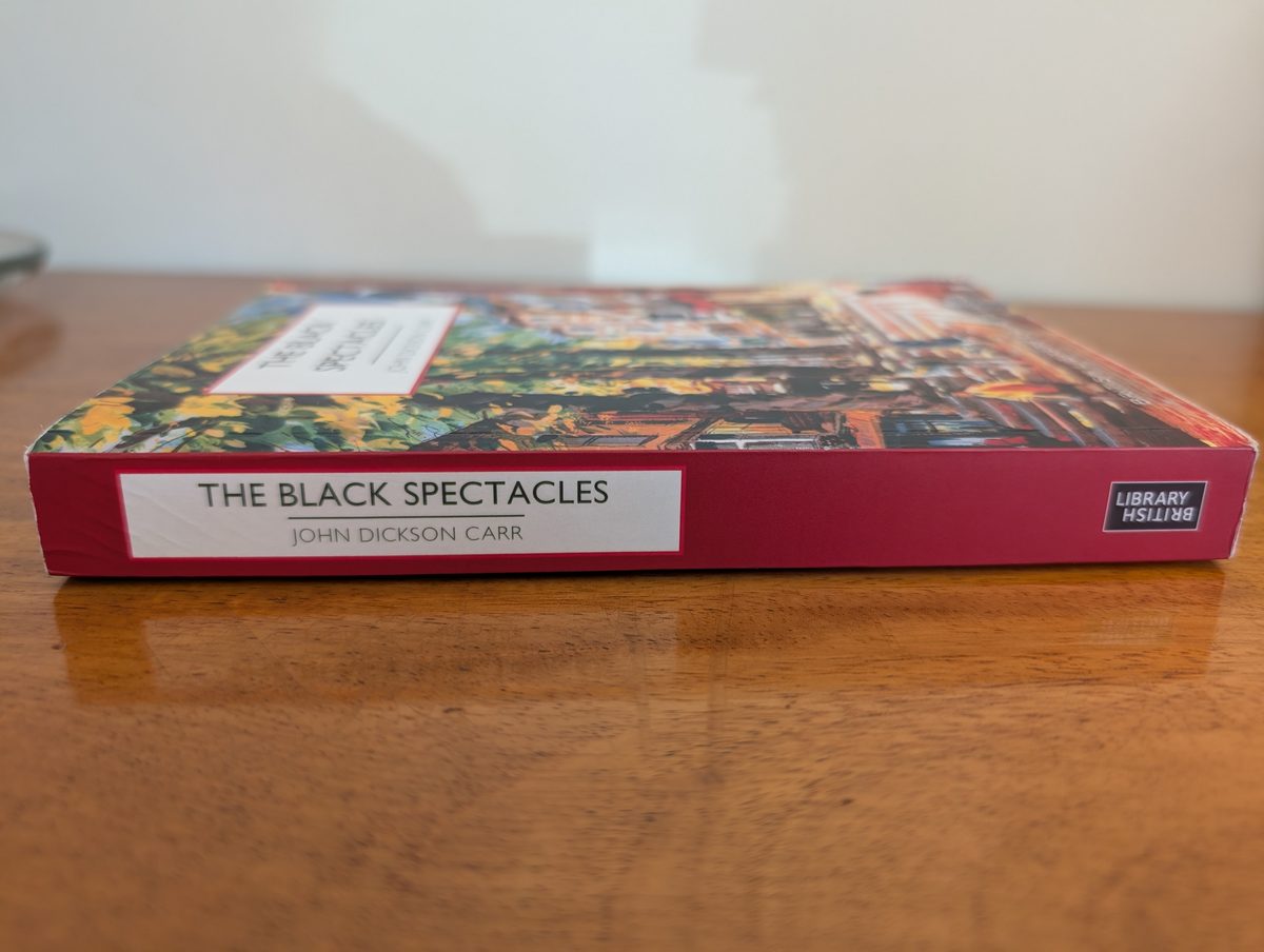 The spine of the 2023 British Library Crime Classics paperback of The Black Spectacles by John Dickson Carr, photographed lying flat. The crimson spine shows a white rectangular panel with 'THE BLACK SPECTACLES' and 'JOHN DICKSON CARR' in dark lettering. The British Library logo appears at one end.