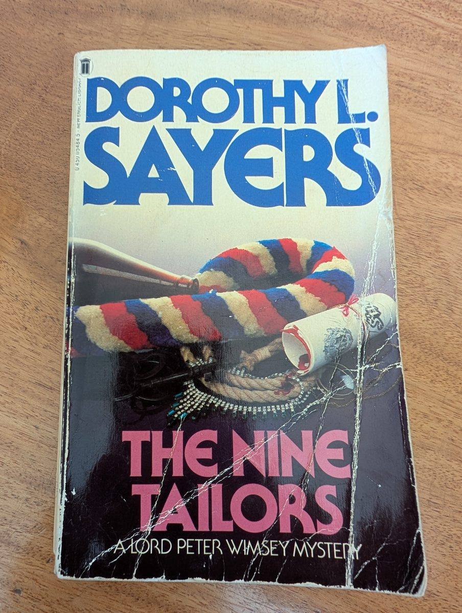 The front cover of the 1982 New English Library paperback edition of The Nine Tailors by Dorothy L. Sayers. The cover shows a dramatic illustration of bell-ringing equipment — a colourful red, blue, white, and yellow striped bell rope and a rolled change-ringing scroll — against a dark background. 'DOROTHY L. SAYERS' appears in blue at the top and 'THE NINE TAILORS / A LORD PETER WIMSEY MYSTERY' in pink lettering below. The cover has significant creasing and surface wear.