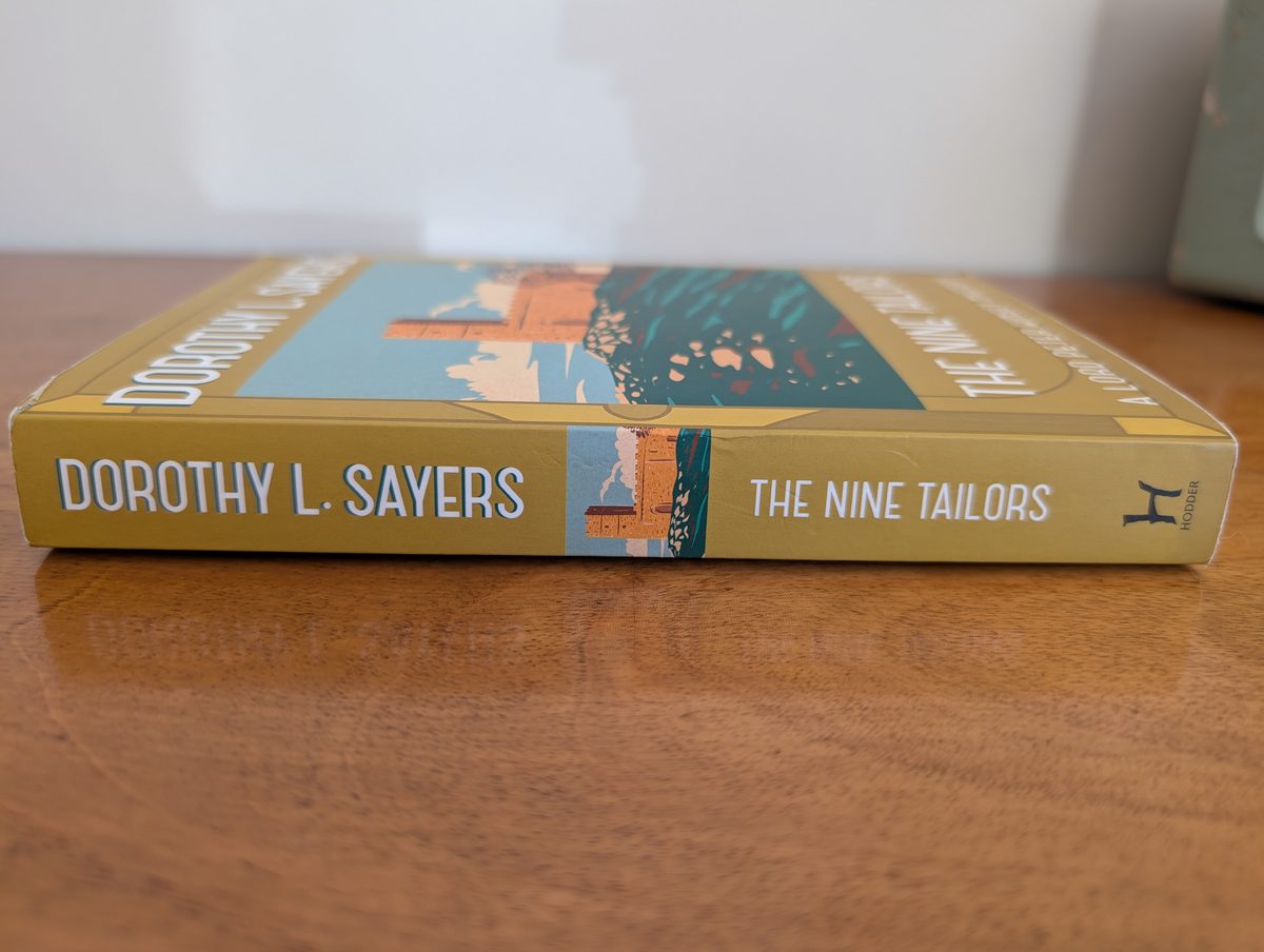 The spine of the 2016 Hodder & Stoughton paperback of The Nine Tailors by Dorothy L. Sayers, photographed lying flat. The mustard-yellow spine shows 'DOROTHY L. SAYERS' and 'THE NINE TAILORS' in white lettering with a small inset of the church tower illustration and the Hodder 'H' logo at one end.